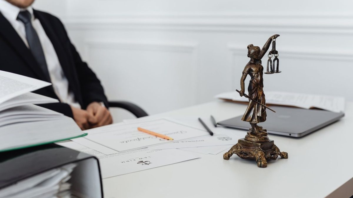 Mesa de trabalho jurídico com documentos, laptop e uma estátua da Justiça ao fundo