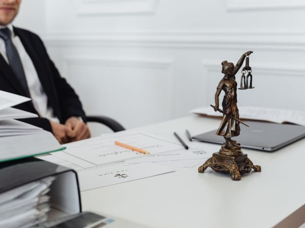 Mesa de trabalho jurídico com documentos, laptop e uma estátua da Justiça ao fundo