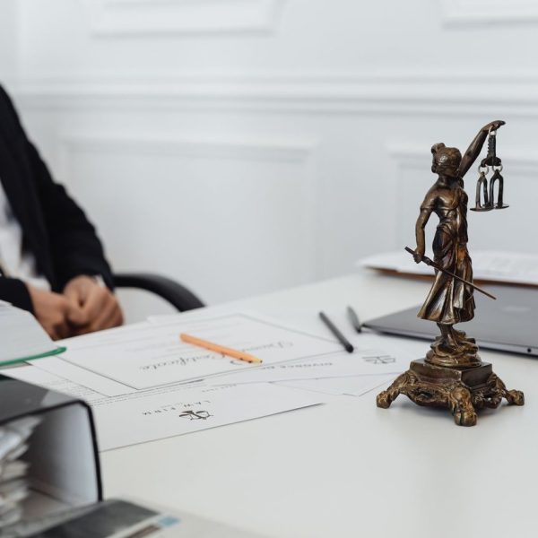 Mesa de trabalho jurídico com documentos, laptop e uma estátua da Justiça ao fundo