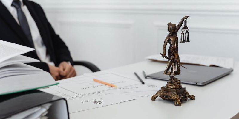 Mesa de trabalho jurídico com documentos, laptop e uma estátua da Justiça ao fundo